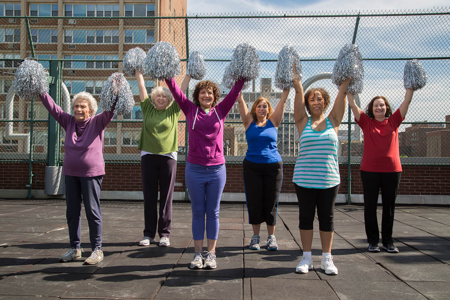 A photograph, exterior on a sunny day - 6 smiling elder women in workout clothes stand in formation on the rooft of a building in a big city, holding silver cheerleader pom-poms over their head, about to cheer.