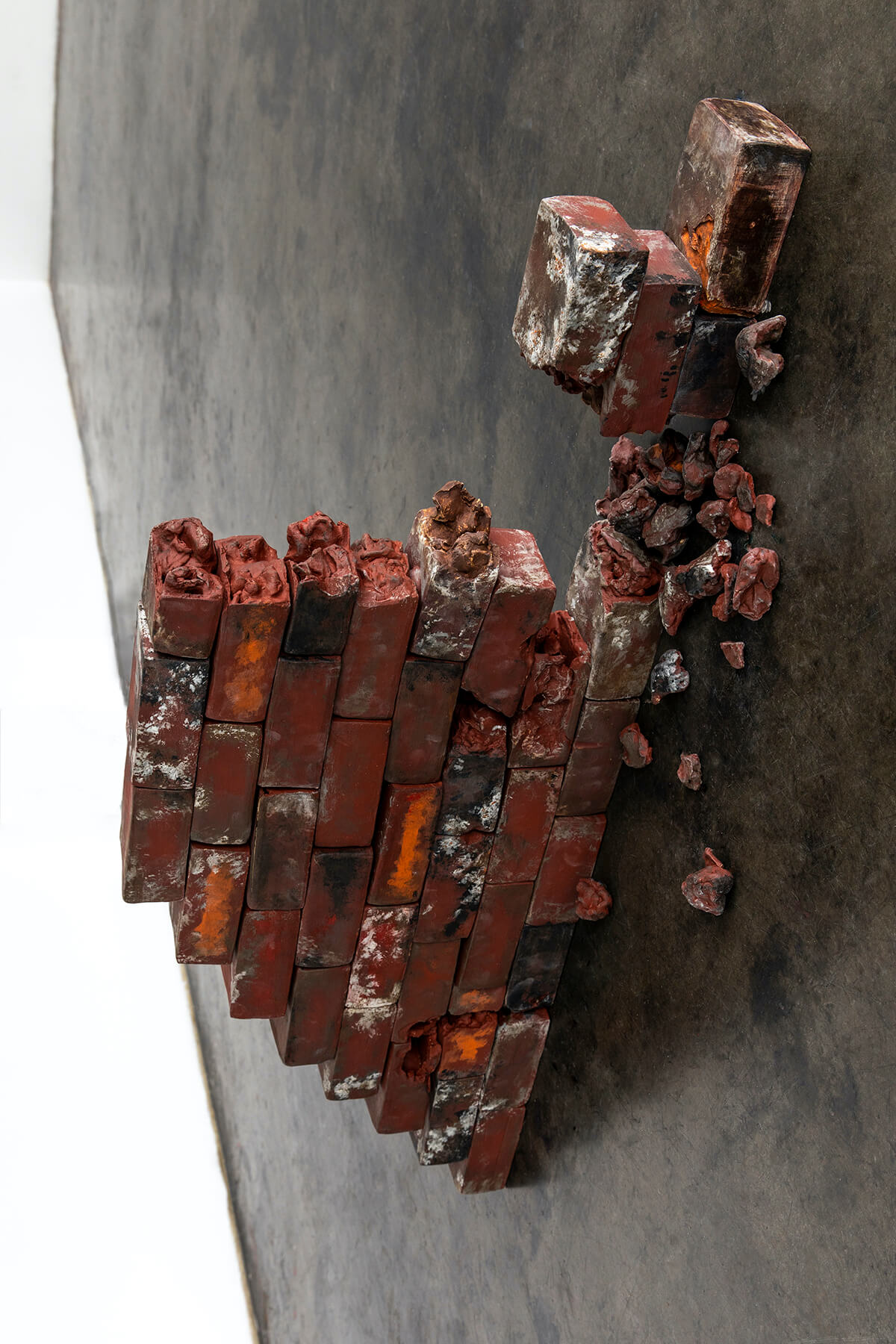 A photograph, interior to an art gallery. On the floor is a stack of rough bricks, mostly dark red but with some white, orange, and black paint. They are arranfed into a small triangular 'wall,' one brick thick, with a portion of the wall missing or collapsed, and on the smooth floor fragments of brick are scattered.