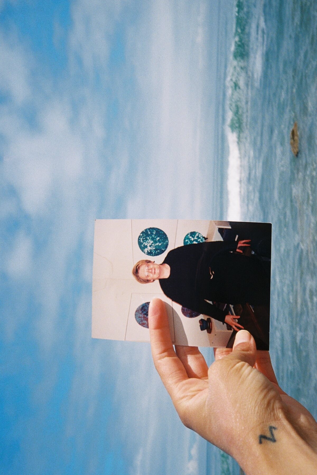 A photograph of a white person's hand holding a photograph of an older white woman. In the background is a large body of water.